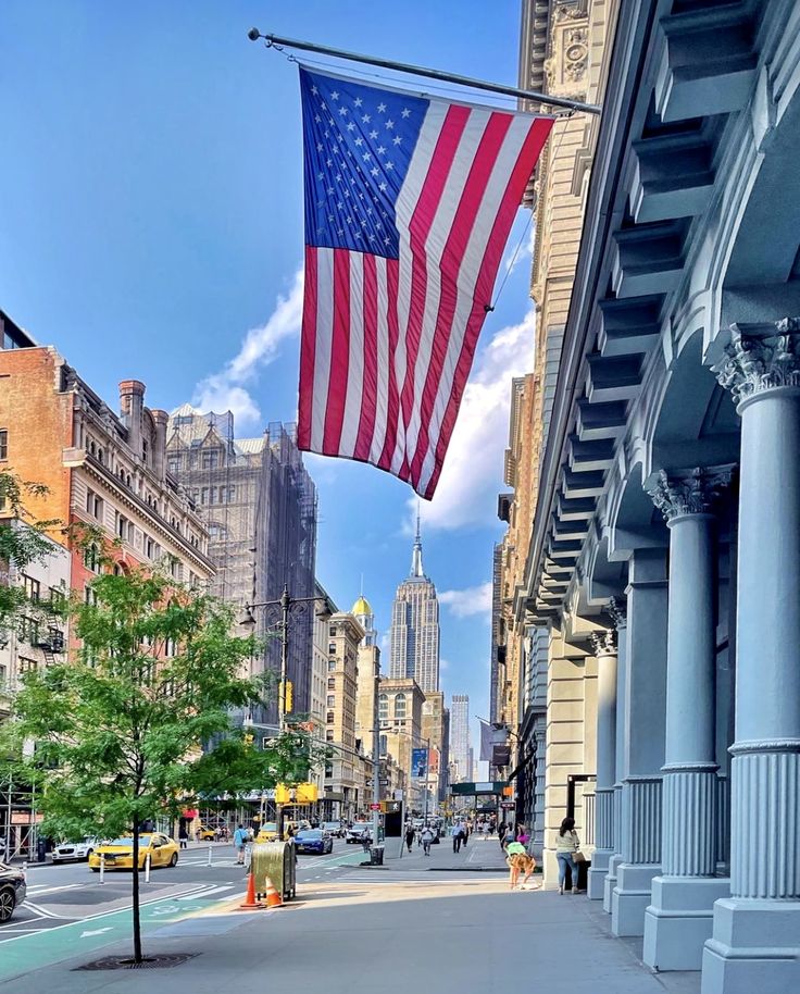 an american flag hanging from the side of a building on a city street with tall buildings in the background