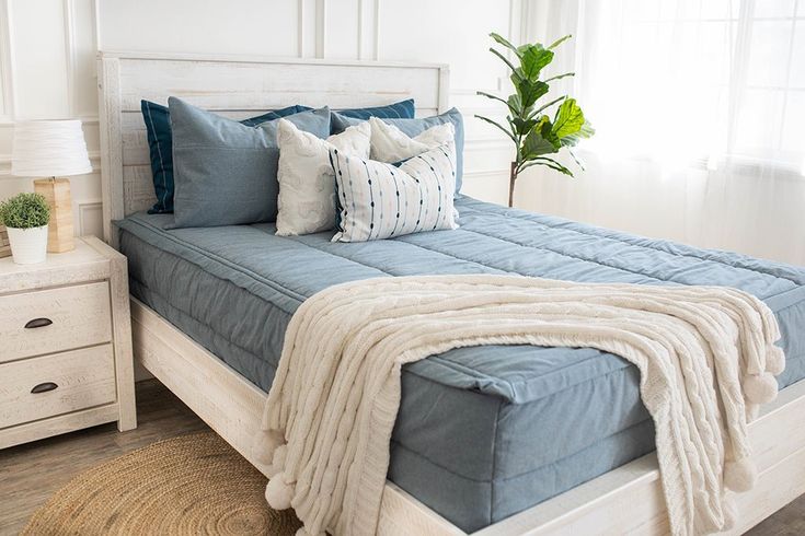 a white bed with blue sheets and pillows in a room next to a plant on a dresser