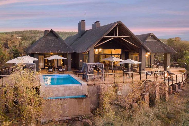 an outdoor swimming pool in front of a house with thatched roof and patio furniture