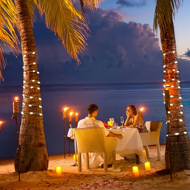 two people sitting at a table on the beach with candles in front of them and palm trees