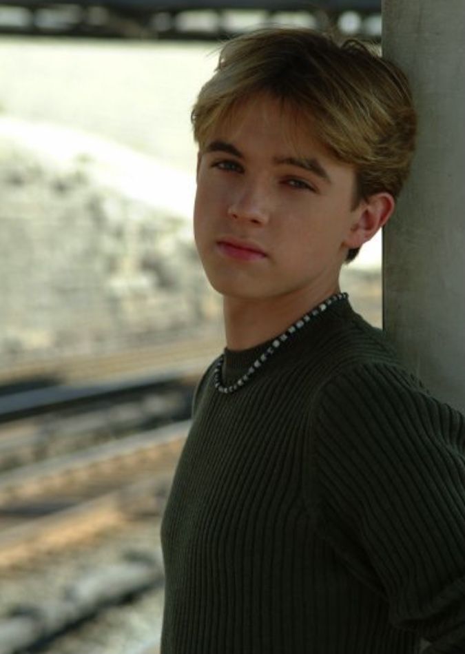 a young man leaning against a pole next to a train track and looking at the camera