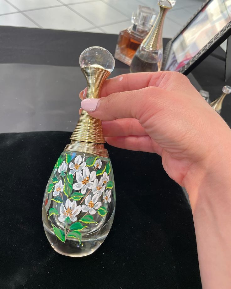 a hand holding a glass bottle with flowers painted on the top and bottom, sitting on a table