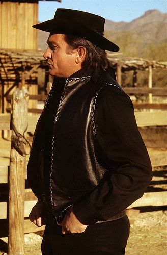 a man wearing a black hat and jacket standing next to a wooden fence in the desert