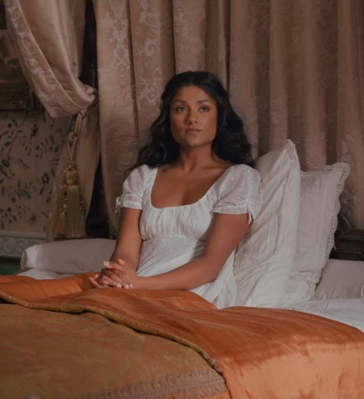 a woman sitting on top of a bed in a white dress and brown bedspread