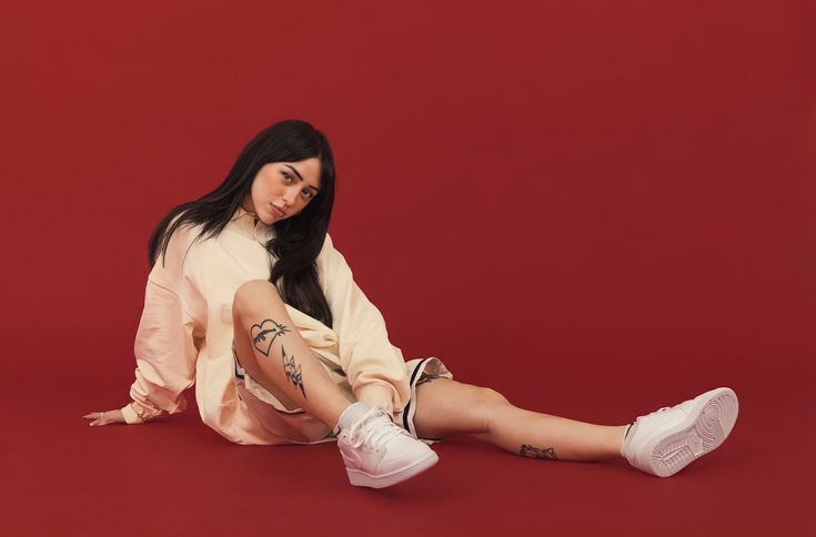 a woman sitting on the floor with her legs crossed and wearing white sneakers, posing for a photo