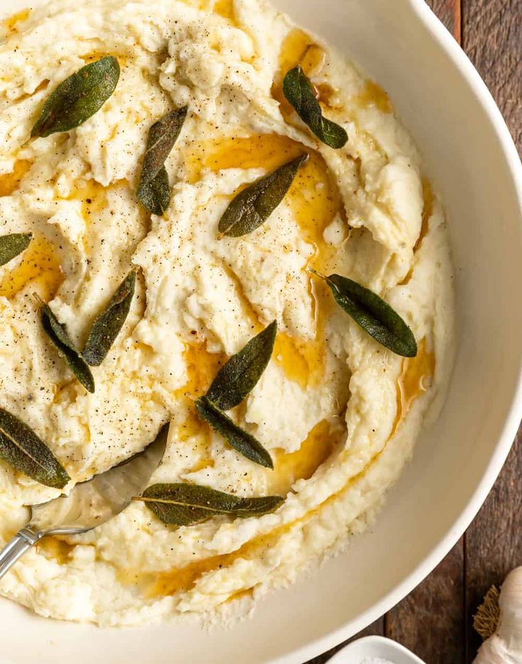 a white bowl filled with mashed potatoes and topped with sage leaves, on top of a wooden table