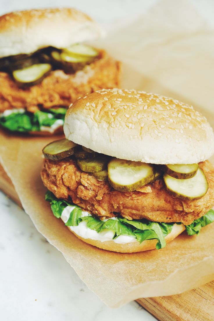 two chicken sandwiches with pickles and cheese on buns sitting on wax paper next to a cutting board