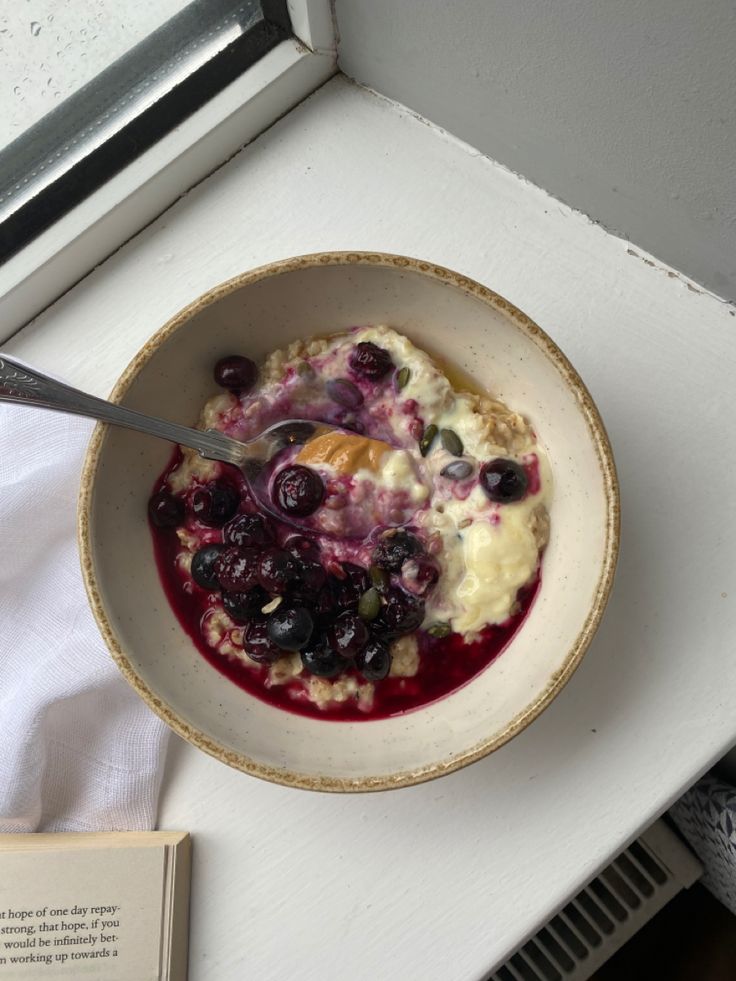 a bowl of oatmeal with blueberries and other toppings sits on a window sill