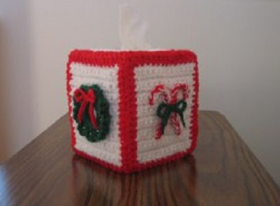 the tissue box is decorated with red and white crocheted ribbon, holly wreaths, and bells