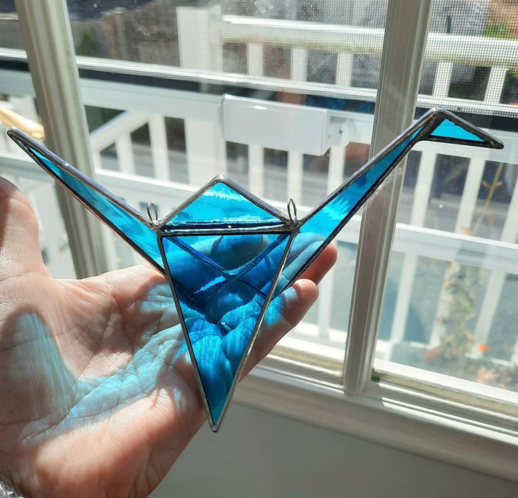 a hand holding a blue glass origami bird in front of a window sill