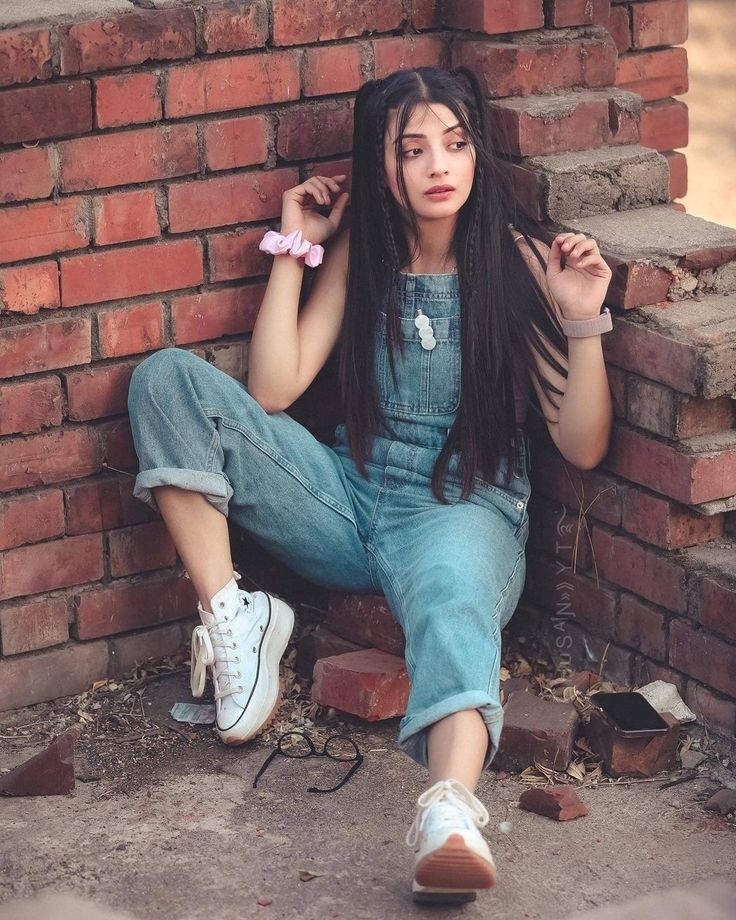 a woman with long hair sitting on the ground next to a brick wall and holding her hands behind her head