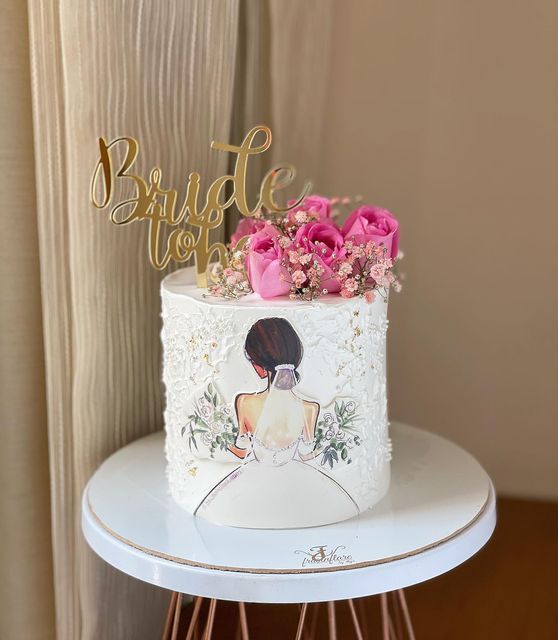 a white wedding cake with pink flowers and a gold name sign on the top that says bride love