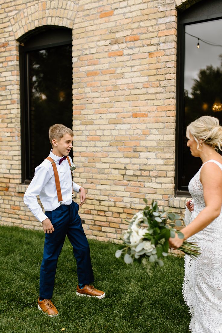 a young boy and woman standing in front of a brick building with flowers on the grass