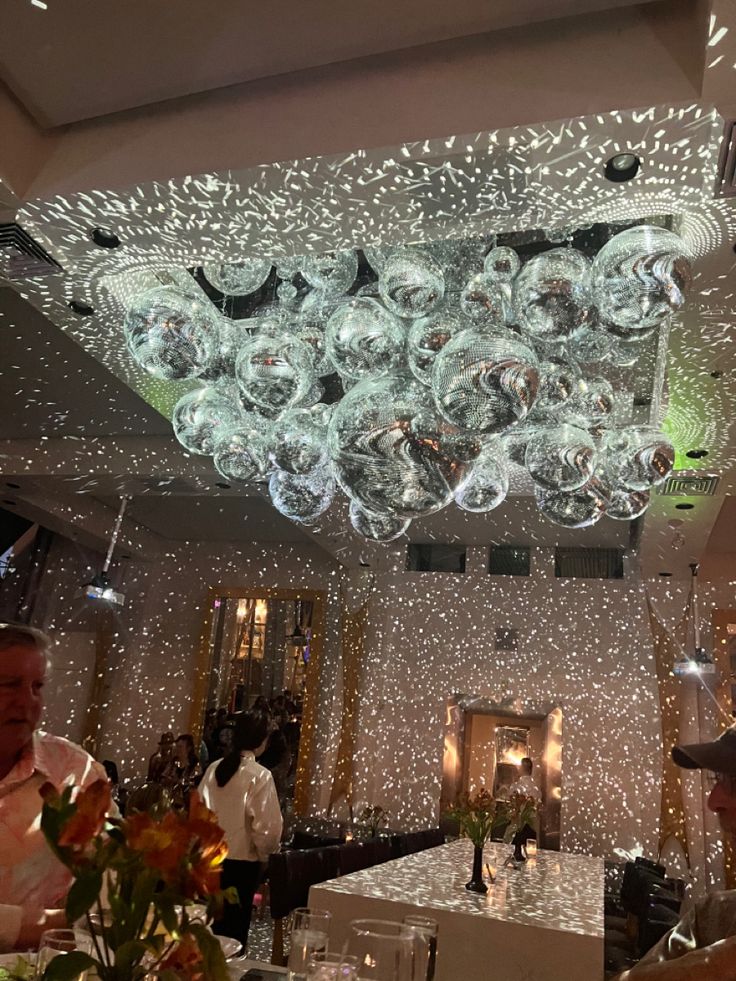 people sitting at tables in a restaurant with lights on the ceiling and chandeliers hanging from the ceiling