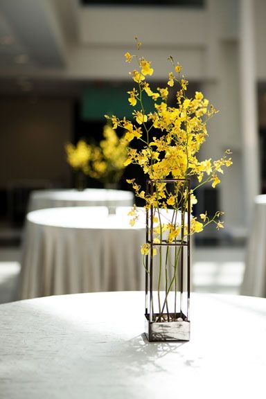 a vase with yellow flowers on top of a white table in a room filled with tables and chairs