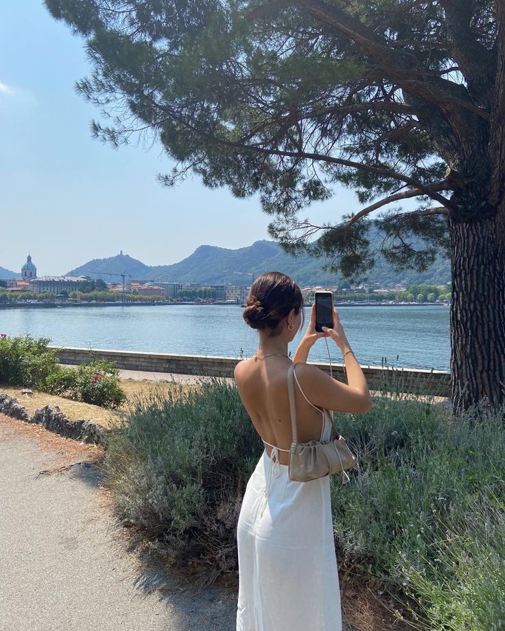 a woman in a white dress taking a photo with her cell phone by the water