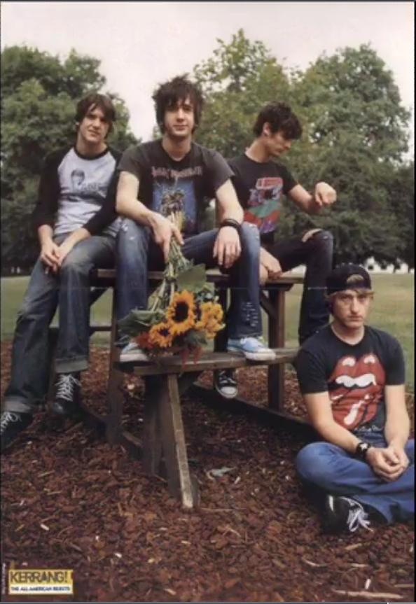 four young men sitting on a bench with sunflowers in the middle and trees behind them