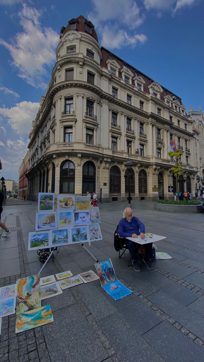 an older man sitting on the ground in front of a building with paintings and drawings