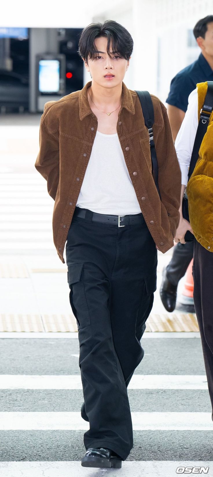 a man walking across a cross walk holding onto a brown jacket over his shoulder and black pants