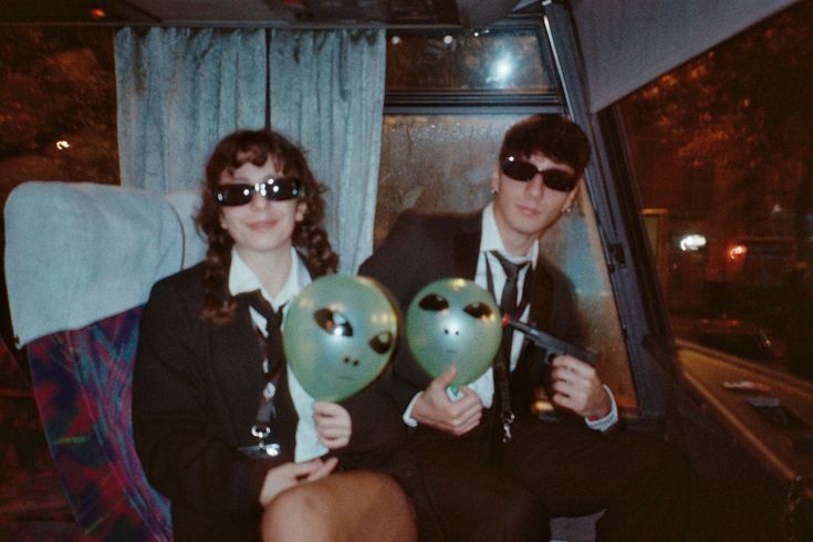 two people sitting on a bus with balloons in front of their faces and wearing sunglasses