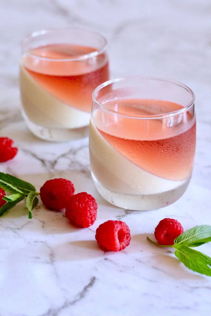 two glasses filled with liquid and raspberries on a marble table