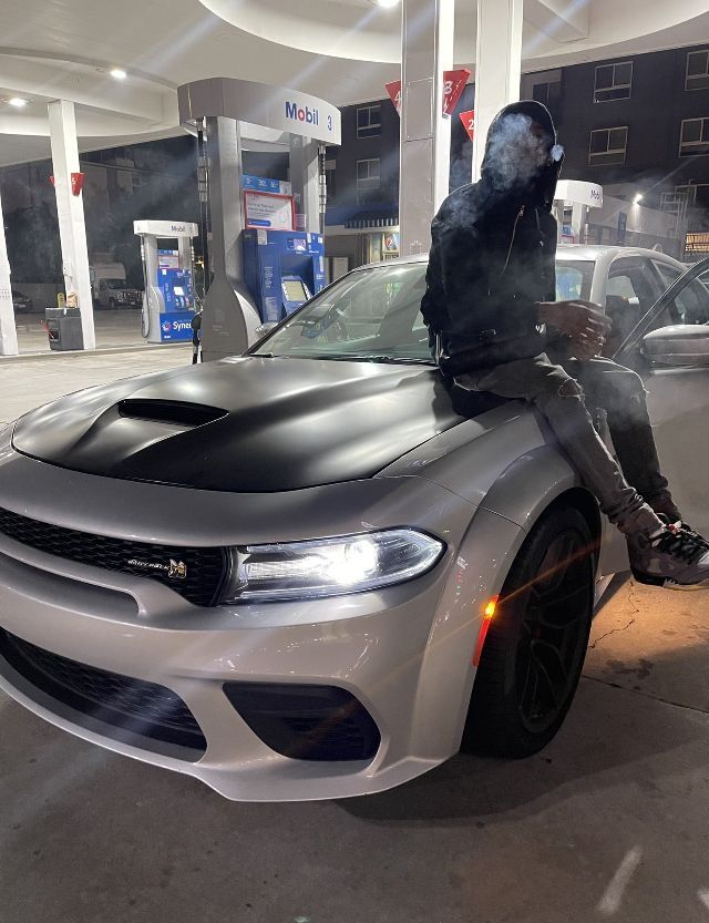 a man sitting on the hood of a silver sports car in a gas station at night