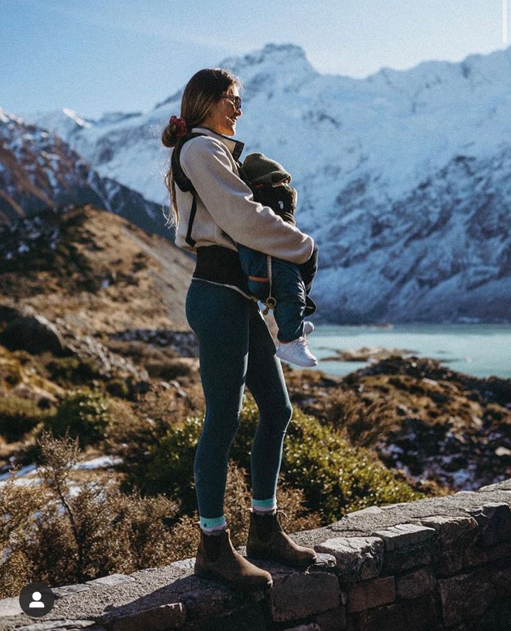 a woman standing on top of a stone wall holding a baby in her arms and looking at the snow covered mountains