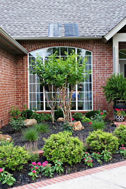 a brick house with lots of plants and flowers in the front yard, along with an arched window