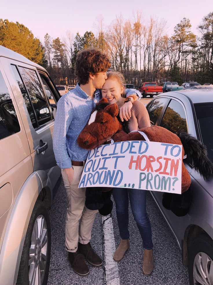 two people standing next to each other in front of a car holding a teddy bear