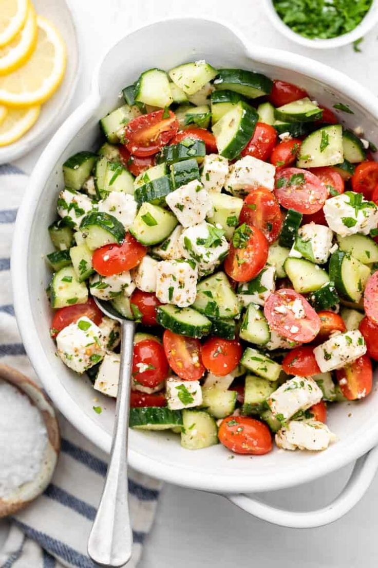 a white bowl filled with cucumber, tomatoes and feta cheese next to lemon slices