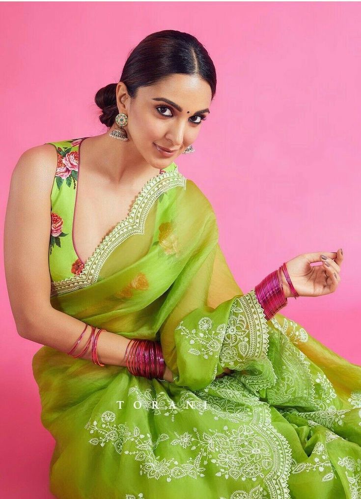 a woman in a green sari sitting on a pink background with her hands clasped