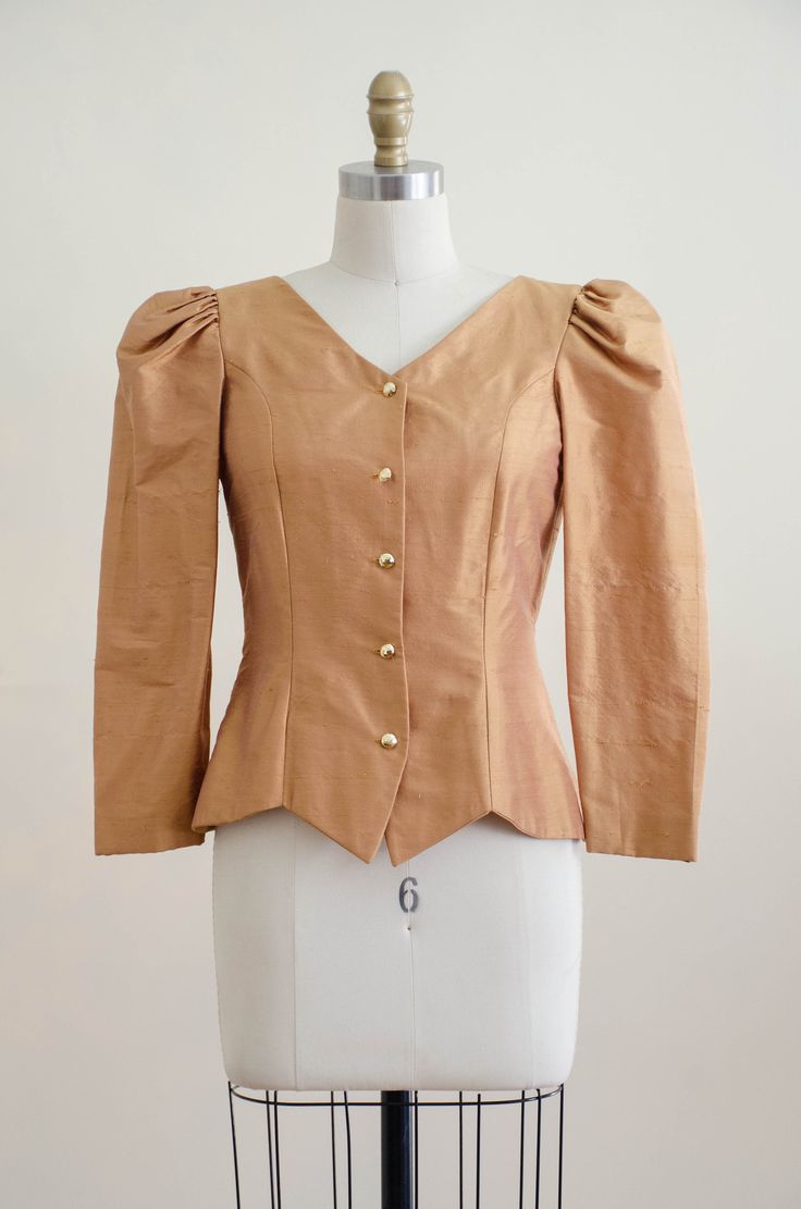 A homemade golden tan jacket, with puffed shoulders, gold faceted buttons, and good tailoring. It is unlined and light enough to be worn as a blouse. ☛ m e a s u r e m e n t s ☚ Bust: 32 Waist: 28 Shoulders: 13.5 Length: 21 ☛ d e t a i l s ☚ Era: 1990s Material: polyester Condition: excellent ☛ v i s i t t h e s h o p ☚ https://etsy.me/2Nd23kg ☛ instagram ┇ poppycockvintage ☛ facebook ┇ poppycockvintage Classic Gold Blouse For Workwear, Fitted Gold Blouse For Office, Fitted Gold Top For Workwear, Fitted Gold Tops For Work, Gold Formal Blouse With Buttons, Gold Blouse For Workwear In Fall, Gold Blouse With Buttons For Work, Fitted Tops With Gold Buttons For Workwear, Gold Blazer With Gold Buttons For Spring