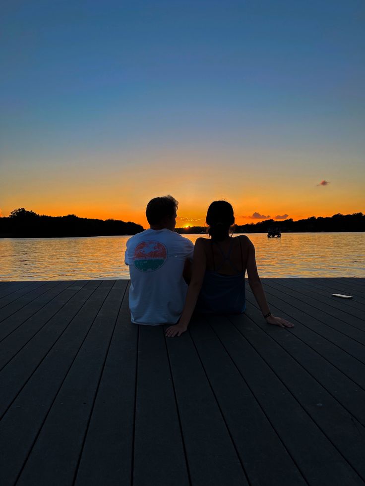 two people sitting on a dock watching the sunset