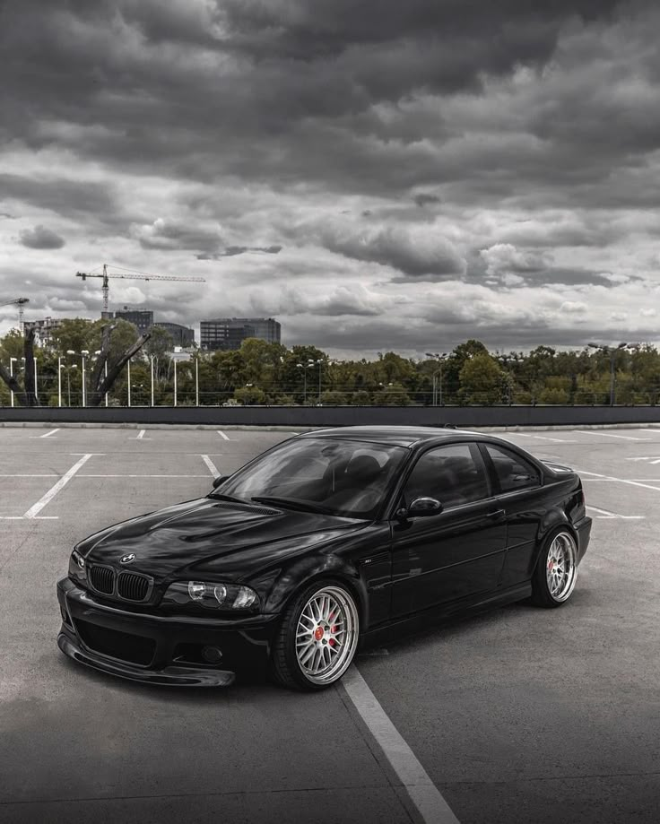 a black car parked in a parking lot with dark clouds above it and trees on the other side