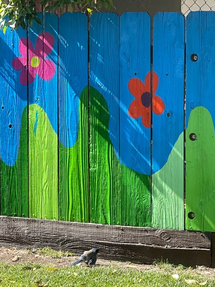 a bird sitting on the ground in front of a fence with flowers painted on it