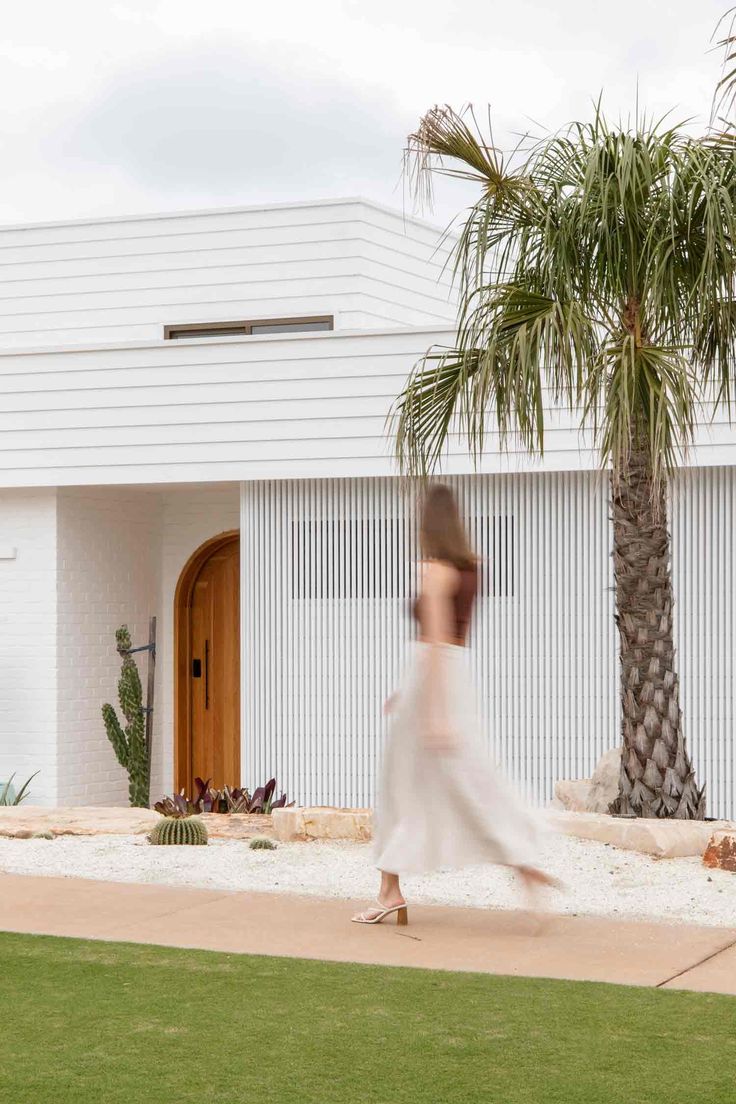 a woman is walking down the sidewalk in front of a white house with palm trees