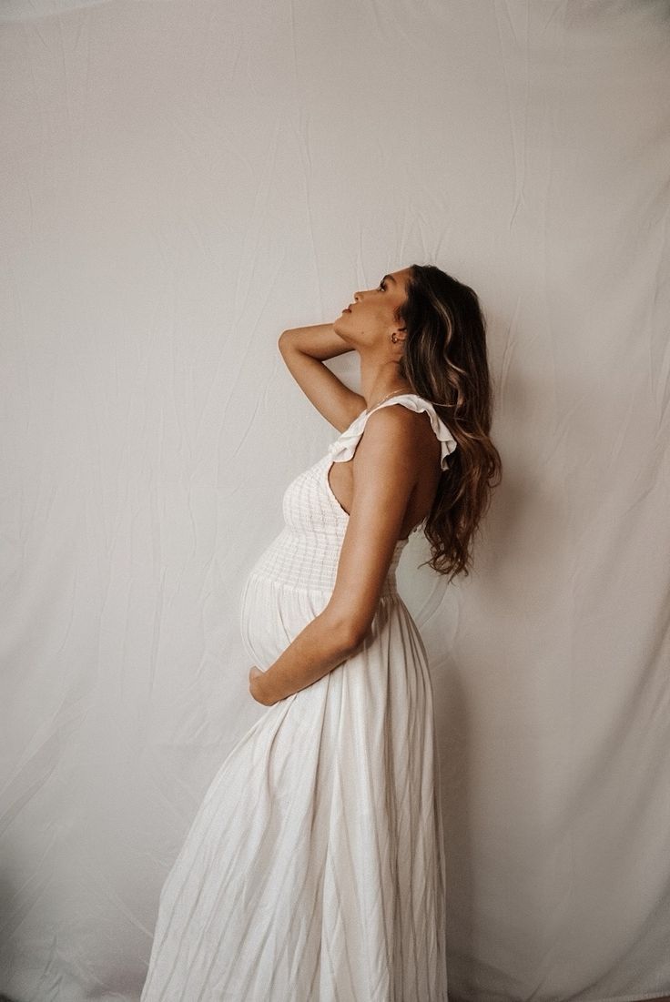 a pregnant woman in a white dress poses for the camera with her hands on her head