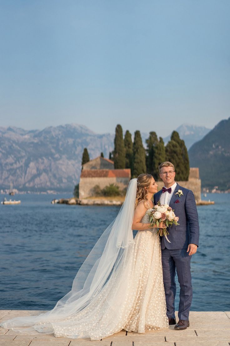 a bride and groom standing on the dock in front of some water with mountains in the background