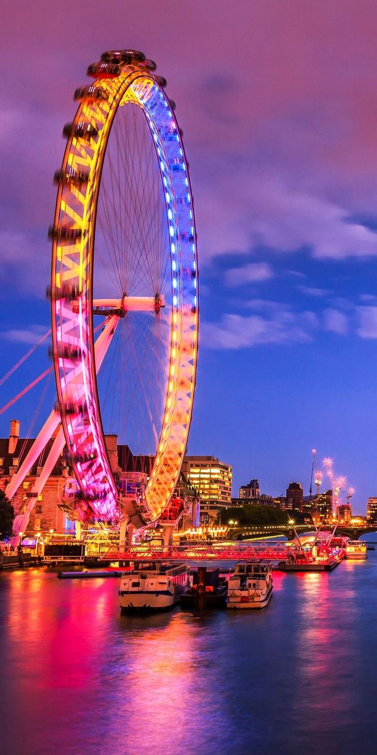 the ferris wheel is lit up at night by the water's edge with boats in the foreground