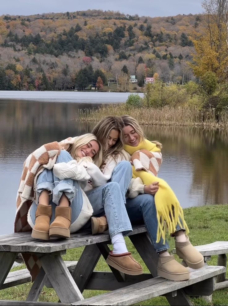 three women are sitting on a picnic table by the water with their arms around each other