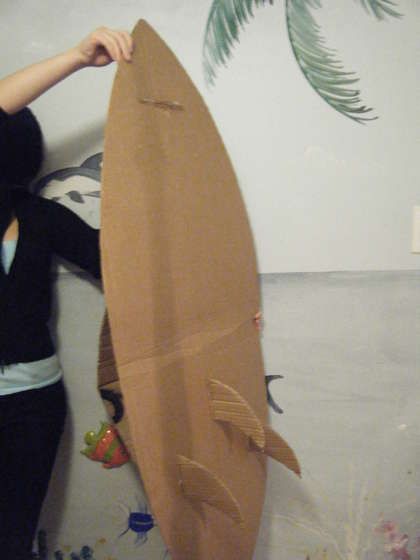 a woman in black shirt holding up a brown surfboard next to wall with palm leaves on it