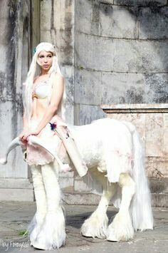 a woman dressed as a white horse standing next to a stone building with an animal on it's back