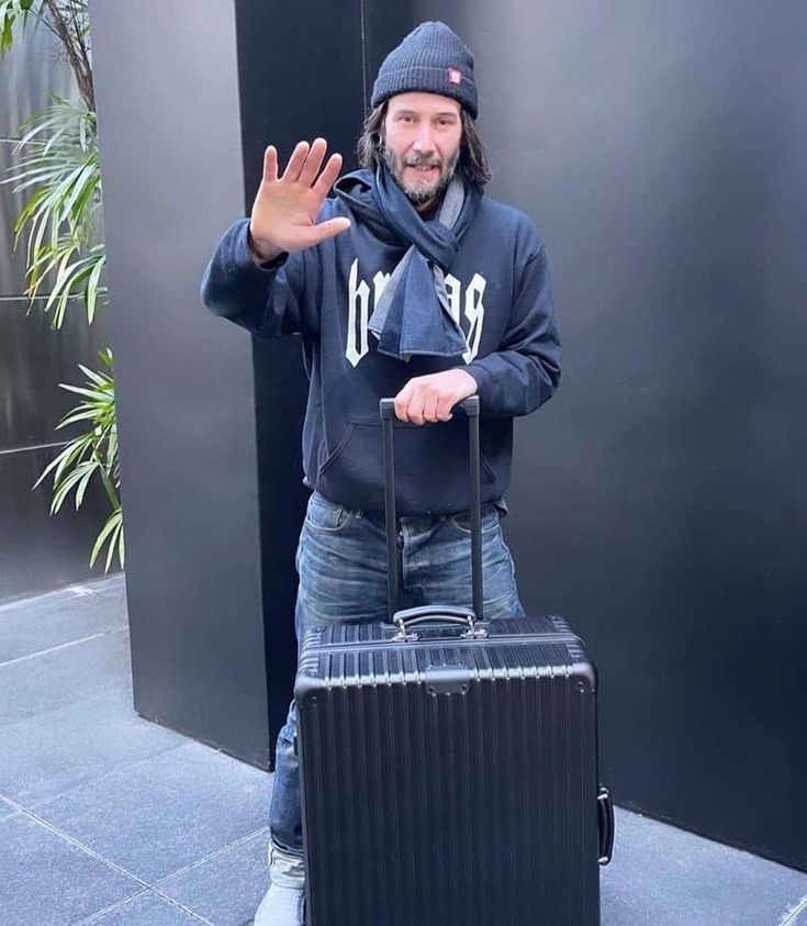 a man is holding his luggage and waving at the camera while wearing a beanie