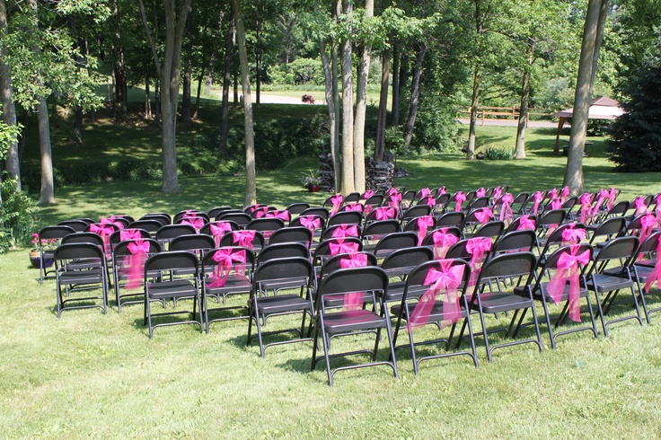 rows of black folding chairs with pink bows on them in front of trees and grass