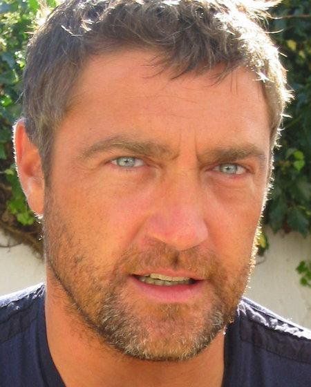 a close up of a person wearing a black shirt and blue eyes with bushes in the background