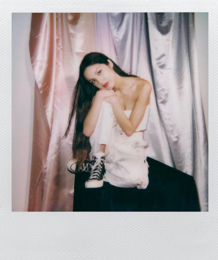 a young woman sitting on top of a bed next to a white curtain with her legs crossed