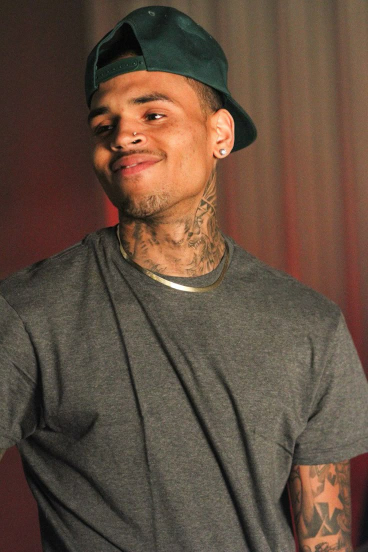 a man with tattoos wearing a hat and looking at the camera while standing in front of a curtain