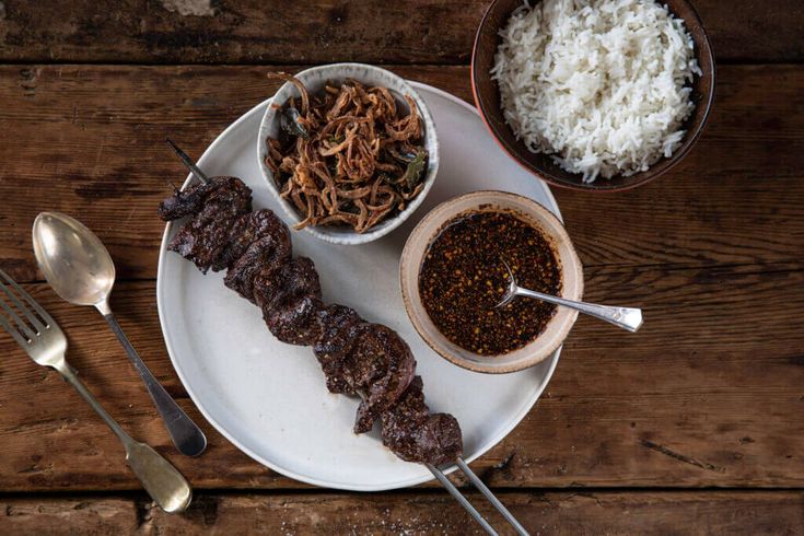 a white plate topped with meat and veggies next to bowls of rice on top of a wooden table