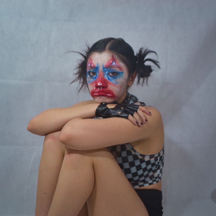 a woman with her face painted like a clown is sitting on the floor and has her arms crossed