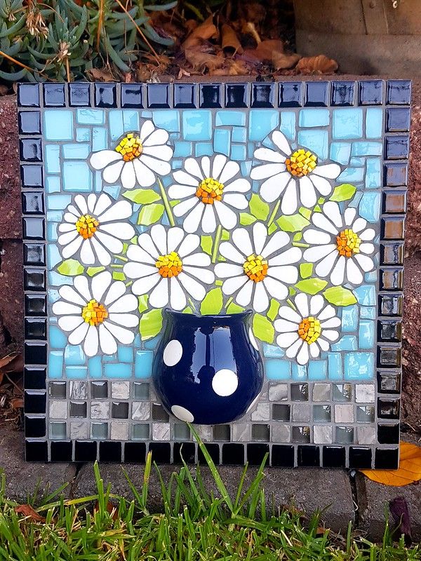 a blue vase with white daisies painted on it sitting in front of a mosaic wall